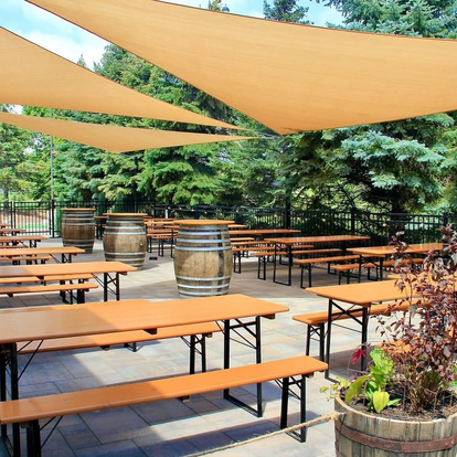 The outdoor area of Elm Creek Brewing features several Classic beer garden sets in pine colour with black frames, arranged under large sun sails. The area is framed by green trees.